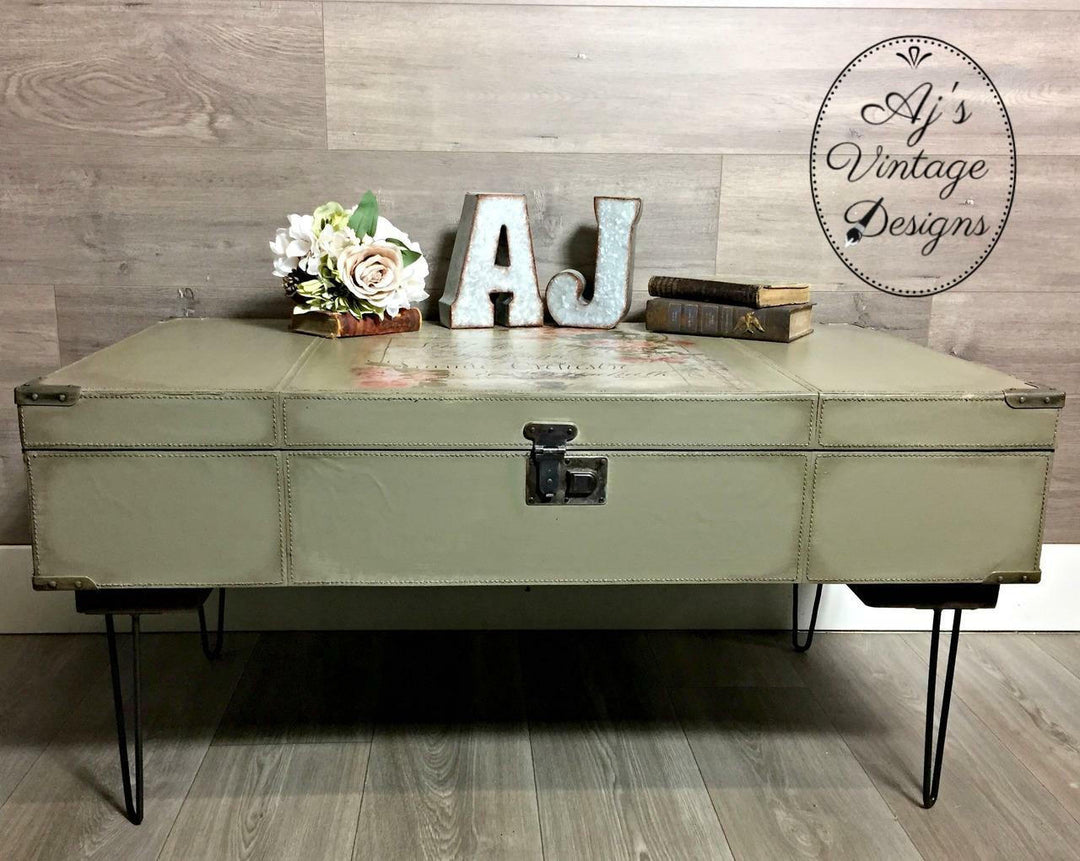 A vintage-style trunk coffee table in a muted sage green color with black hairpin legs, styled with decorative letters, books, and flowers against a wood-paneled wall. A circular logo for 'Aj's Vintage Designs' is visible in the top right corner.