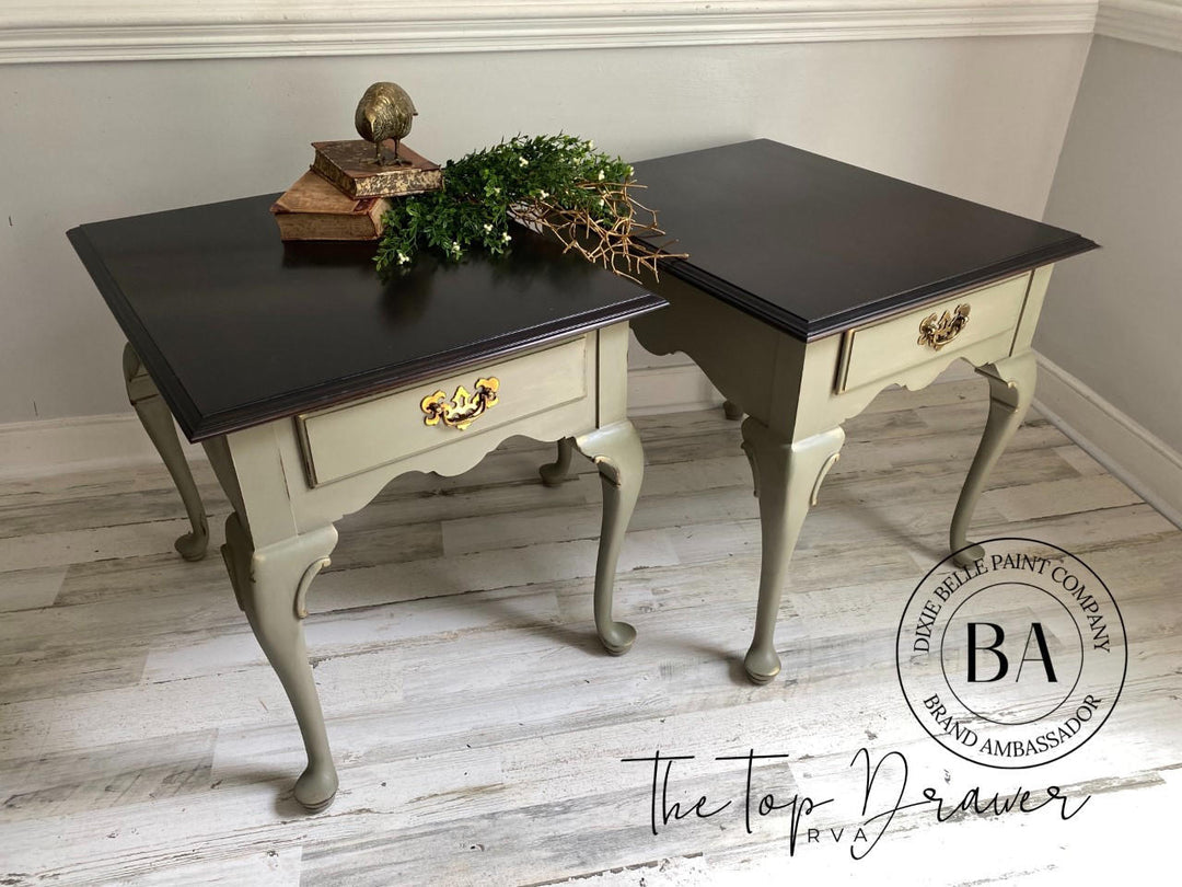 A pair of vintage-style writing desks in a distressed sage green finish with dark brown tops and gold hardware, styled on a light wood floor with decorative greenery on top.