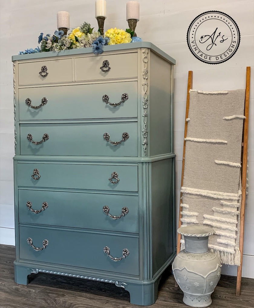 A vintage-style dresser with a vertical ombre transition from cream at the top to a muted sage green at the bottom, styled in a home setting with candles, flowers, a decorative vase, and a wall hanging.