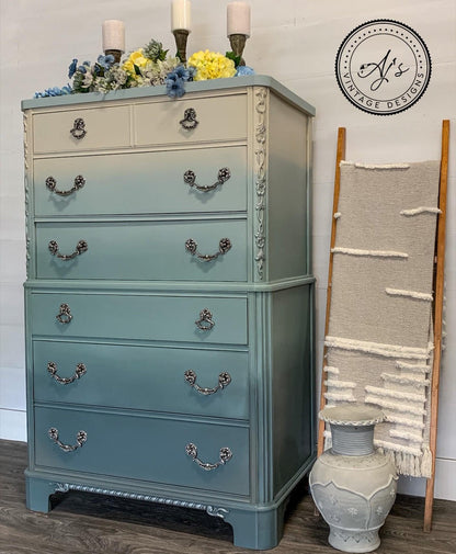A vintage-style dresser with a vertical ombre transition from cream at the top to a muted sage green at the bottom, styled in a home setting with candles, flowers, a decorative vase, and a wall hanging.