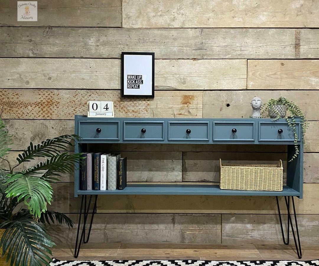 A blue console table with five drawers and an open lower shelf, styled against a rustic wood plank wall with books, a basket, a small statue, and a framed print.