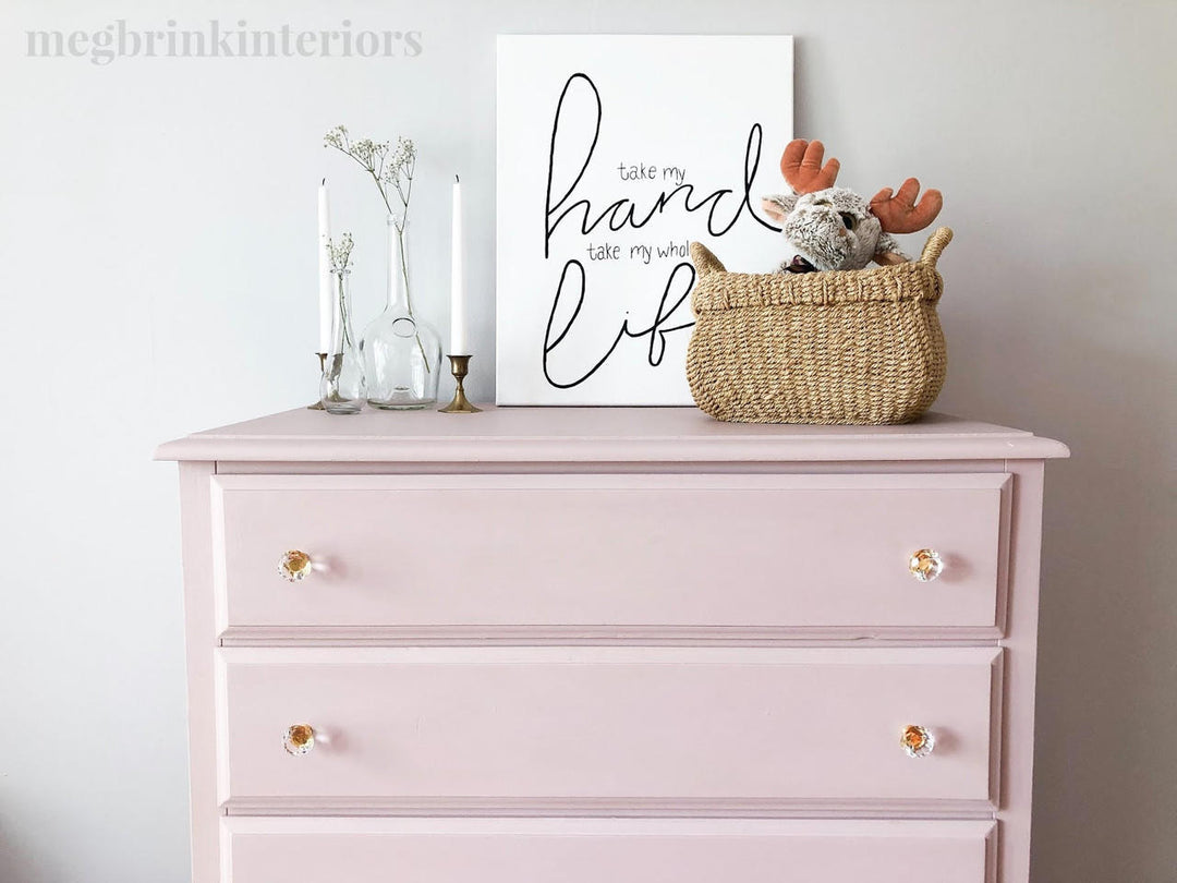 A light pink three-drawer dresser against a white wall, styled with a white sign that reads 'take my hard work take my life,' a woven basket with a plush toy and orange reindeer ears, and two white taper candles in glass holders.