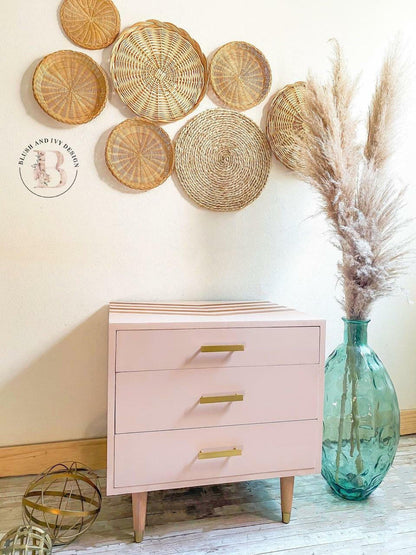 A light pink three-drawer nightstand with gold metal handles and light wood legs, styled in a room setting with a wall of woven baskets, a large teal glass vase with pampas grass, and a small decorative sphere on the floor.