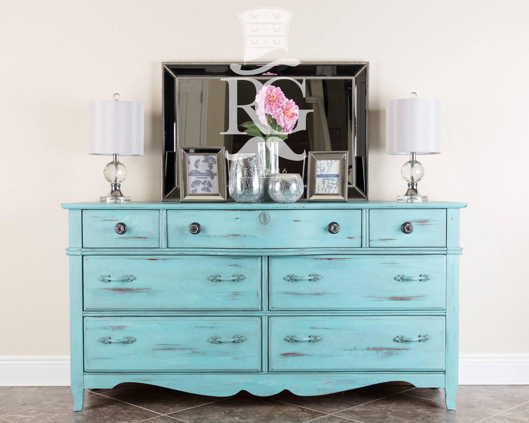 A distressed turquoise blue wooden dresser with six drawers, styled with a large black-framed mirror, two table lamps, and decorative frames against a beige wall.