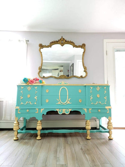 A turquoise and gold painted console table with an ornate gold mirror above it, set in a bright room with light gray walls and wood floors.