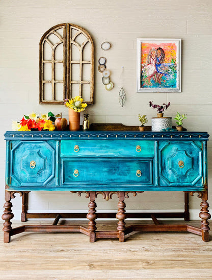 A turquoise and teal distressed wooden console table with carved legs, styled with autumn decorations, potted plants, and wall art in a rustic room setting.