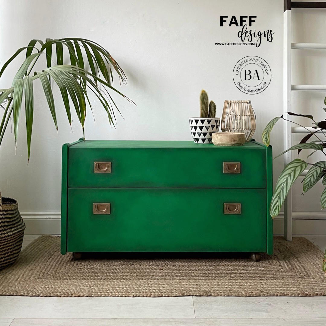 A bright green two-drawer chest of drawers with brass handles, styled in a room setting on a woven rug with house plants and decorative items on top. The Faff Designs logo is visible in the top right corner.