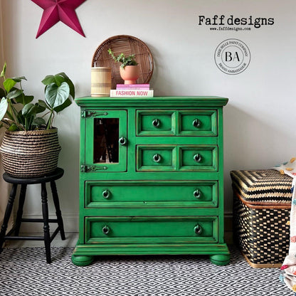 A bright green painted wooden chest of drawers with distressed detailing, shown in a styled room setting with a potted plant on a stool, a basket, and patterned rug. The image includes 'Faff designs' branding and a circular BA logo watermark.