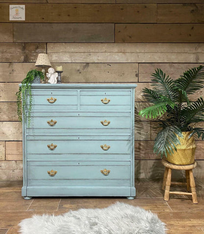 A distressed light blue painted wooden chest of drawers with brass handles, styled in a rustic room with a wood plank wall, a potted palm on a stool, and a white faux fur rug.