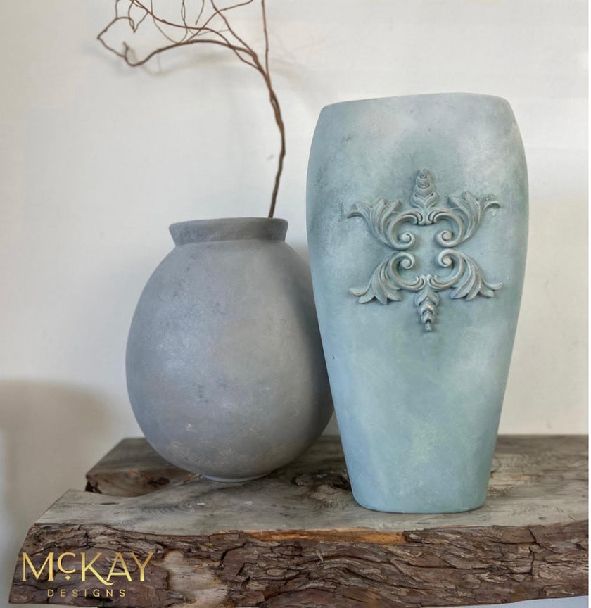 Two matte ceramic vases in a dusty blue-gray finish, displayed on a rustic wood slice against a plain off-white wall. The larger vase on the right features a raised baroque-style scrollwork motif, while the smaller vase on the left is plain.