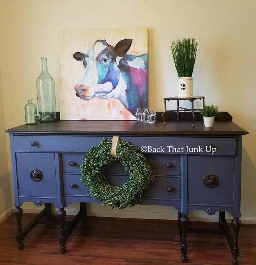 Blue dresser with a wreath, painting of a cow, and decorative items against a beige wall.