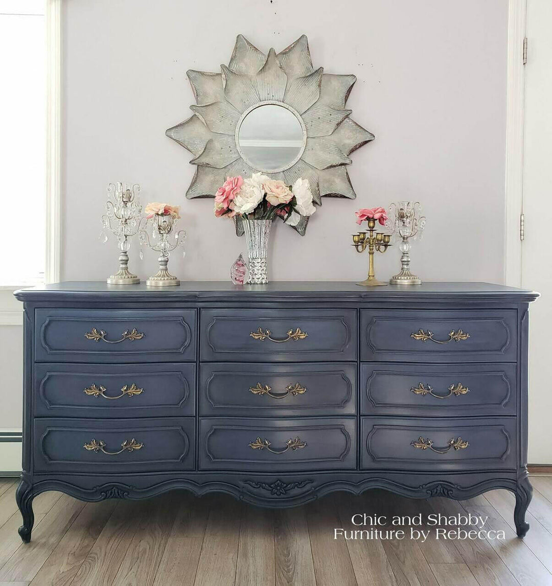 Navy blue dresser with decorative items in a room setting, featuring a large mirror and flowers.