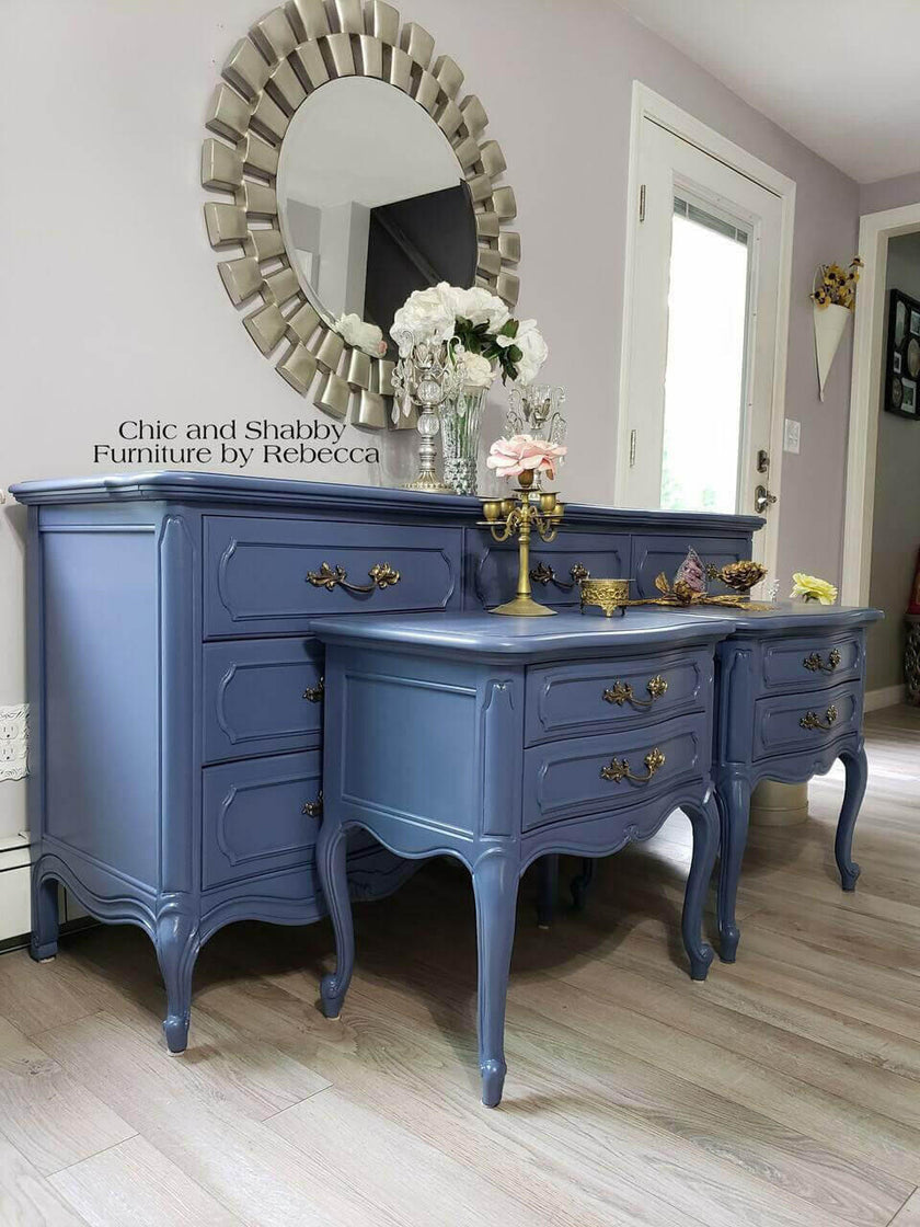 A painted blue vanity desk and matching side cabinet set with ornate brass handles, styled in a room with a large sunburst mirror, white flowers, and light wood floors. Text on the image reads 'Chic and Shabby Furniture by Rebecca'.