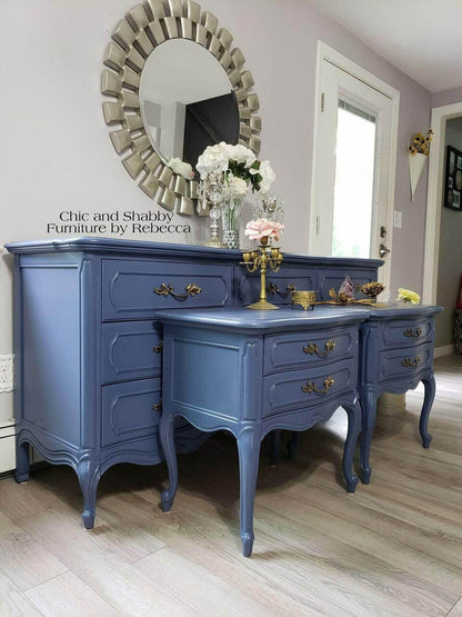 A painted blue vanity desk and matching side cabinet set with ornate brass handles, styled in a room with a large sunburst mirror, white flowers, and light wood floors. Text on the image reads 'Chic and Shabby Furniture by Rebecca'.