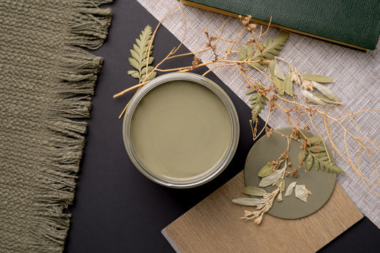 An open round metal paint can filled with muted sage green paint, styled on a dark tabletop with dried leaves, a textured olive green fringed fabric, a woven rug, a wooden board, and a green book.