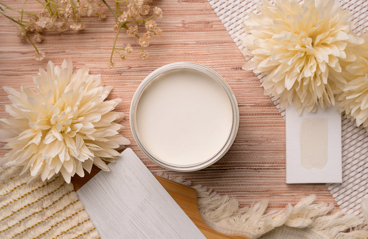 An open round jar of cream-colored Magnolia paint or pigment sits on a woven mat, surrounded by large pale flowers, a paint swatch card, and a wooden palette knife.