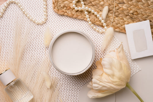 A round gardenia paint jar, styled in a flat lay with a pearl necklace, woven mat, dried grass, a white tulip, and a small card on a light neutral background.