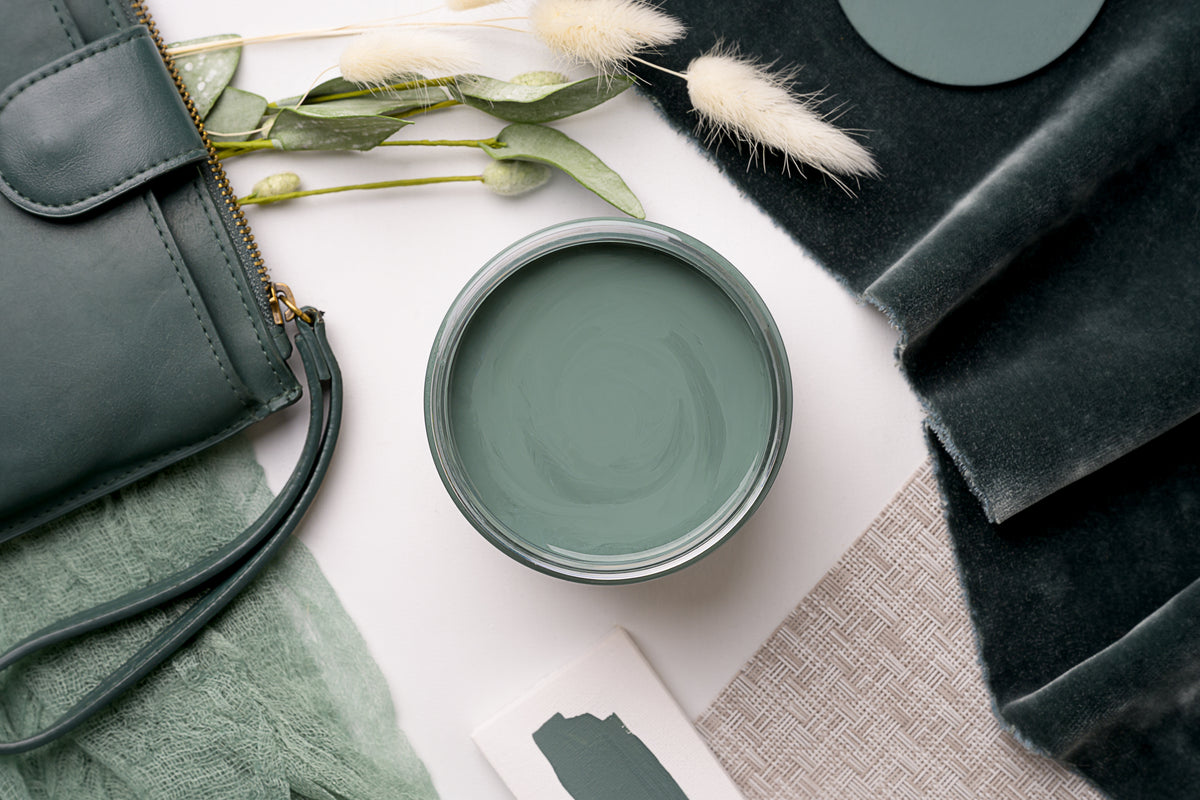 An open round paint can filled with muted sage green paint, styled in a flat lay with a dark green handbag, velvet fabric, dried bunny tail grass, and a small paint swatch card on a white surface.