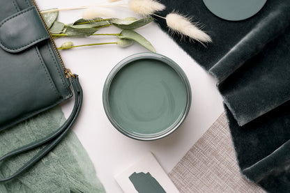 An open round paint can filled with muted sage green paint, styled in a flat lay with a dark green handbag, velvet fabric, dried bunny tail grass, and a small paint swatch card on a white surface.
