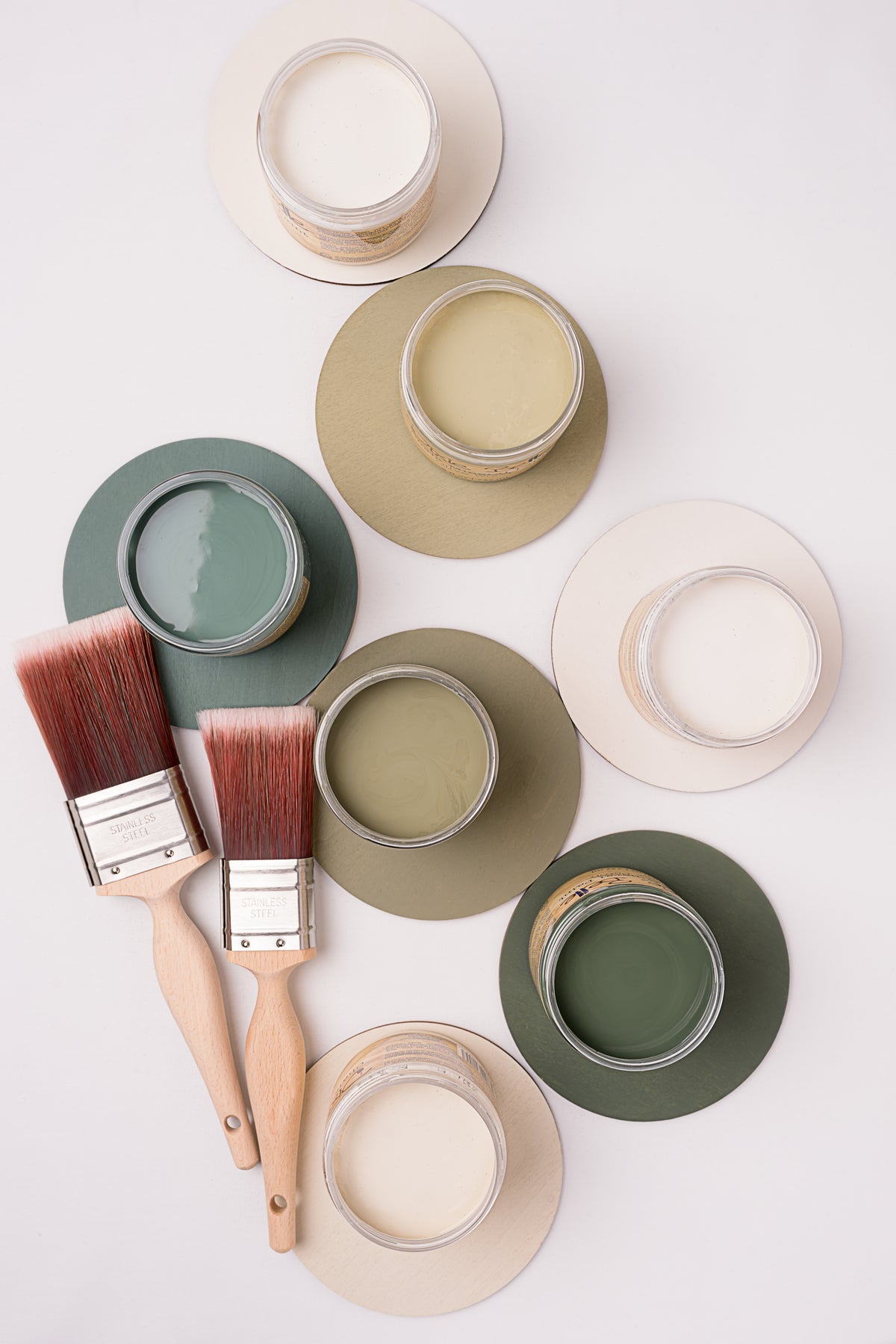 A styled flat-lay of paint swatches and paintbrushes on a white background. Several round paint cans with cream, sage green, and olive paint are arranged on matching circular swatches, alongside two wooden-handled paintbrushes with reddish-brown bristles.