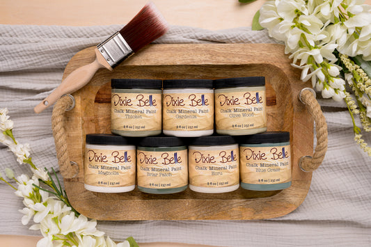 Set of Dixie Belle Chalk Mineral Paint jars on a wooden tray with a brush, surrounded by flowers.