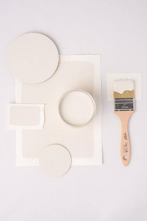 A flat-lay of paint swatches and a paintbrush on a light gray background. The set includes several circular and rectangular swatches in warm off-white and cream tones, a small round tin of cream-colored paint, and a wooden-handled paintbrush with cream paint on the bristles.