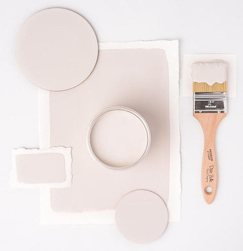 A flat-lay of a paint color palette featuring a light blush pink paint swatch, matching paper circles and swatches, a small tin of paint, and a wooden paint brush with a light pink brush stroke on the bristles, all arranged on a white background.