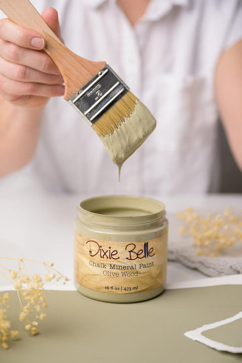 Person using a brush to apply Olive Wood color from a Pixie Belle jar on a neutral background