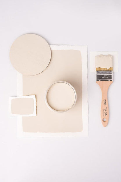 A flat-lay of paint color testing materials on a white background, including a round paint swatch, a rectangular paint swatch, an open jar of cream-colored paint, and a wooden-handled paintbrush.