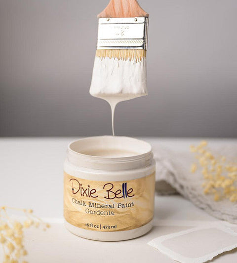 A jar of Dixie Belle Chalk Mineral Paint in the color Gardenia, with a paintbrush lifting white paint above the open jar against a neutral gray background.
