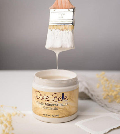 A jar of Dixie Belle Chalk Mineral Paint in the color Gardenia, with a paintbrush lifting white paint above the open jar against a neutral gray background.