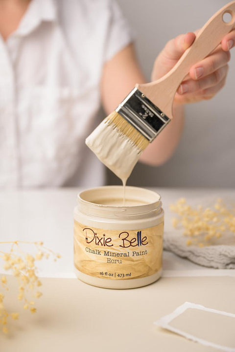 A person in a white shirt holds a paintbrush dipped in thick beige paint above an open jar of Dixie Belle Chalk Mineral Paint. The scene is softly lit with blurred dried flowers and a neutral background.