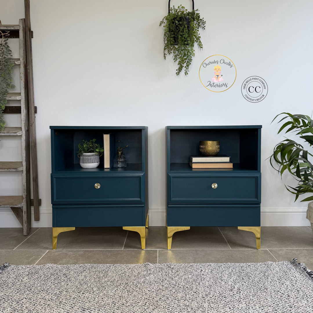 Two dark blue nightstands with gold feet in a room with plants and a ladder.