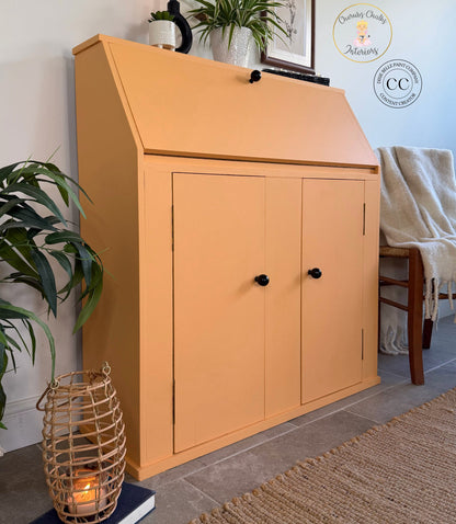 A peach-colored wooden writing desk cabinet with a slanted top and two doors, styled in a home interior next to a chair, plants, and a woven basket candle holder.