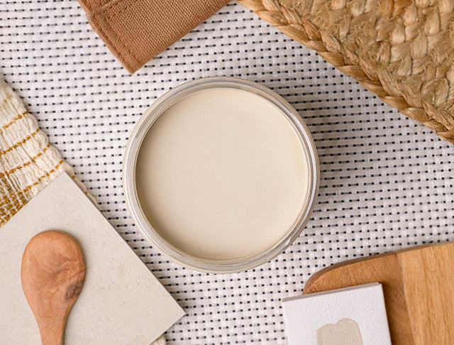 An open round jar filled with a smooth off-white paint, placed on a white woven mat with a wooden spoon, cloth, and other neutral props around it.