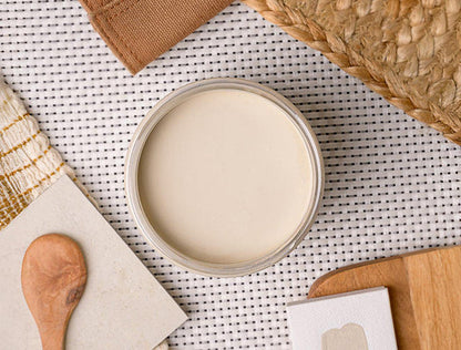 An open round jar filled with a smooth off-white paint, placed on a white woven mat with a wooden spoon, cloth, and other neutral props around it.