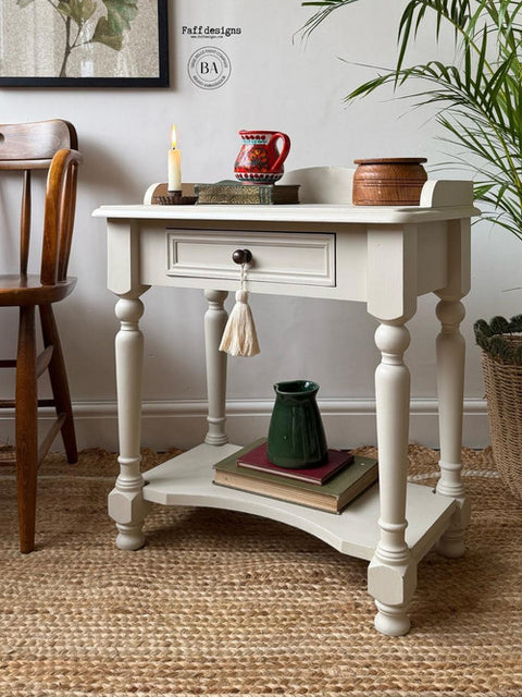 A distressed white wooden console table with a single drawer, turned legs, and a lower shelf, styled in a home interior with books, a green vase, and decorative items.