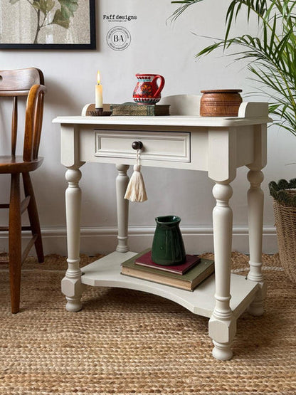 A distressed white wooden console table with a single drawer, turned legs, and a lower shelf, styled in a home interior with books, a green vase, and decorative items.