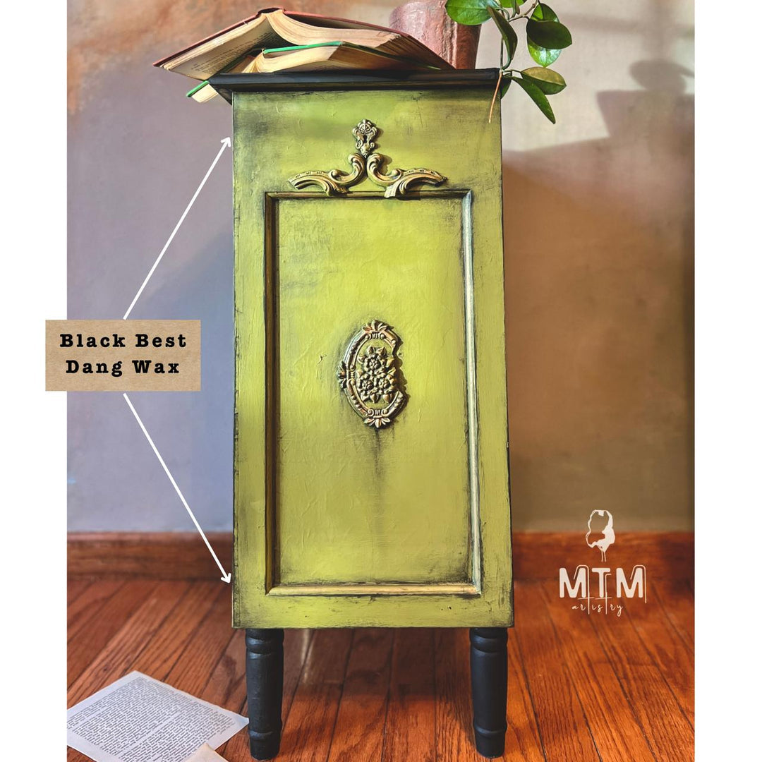 A vintage-style nightstand or cabinet with a distressed olive green finish and black legs, shown in a room setting with a wooden floor and wall. Text on the image indicates the finish is 'Black Best Dang Wax', and there is an 'MTM staging' logo watermark.