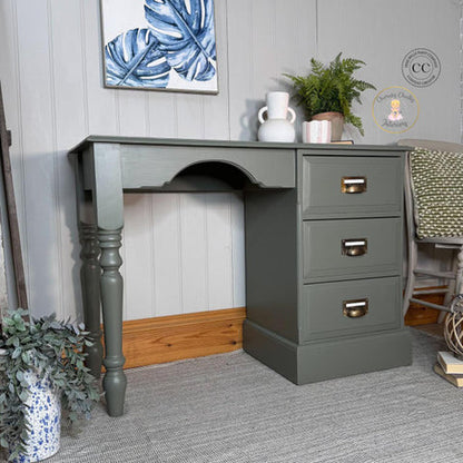 A painted wooden writing desk with three drawers and turned legs, shown in a styled room setting with wall decor, plants, and a chair.