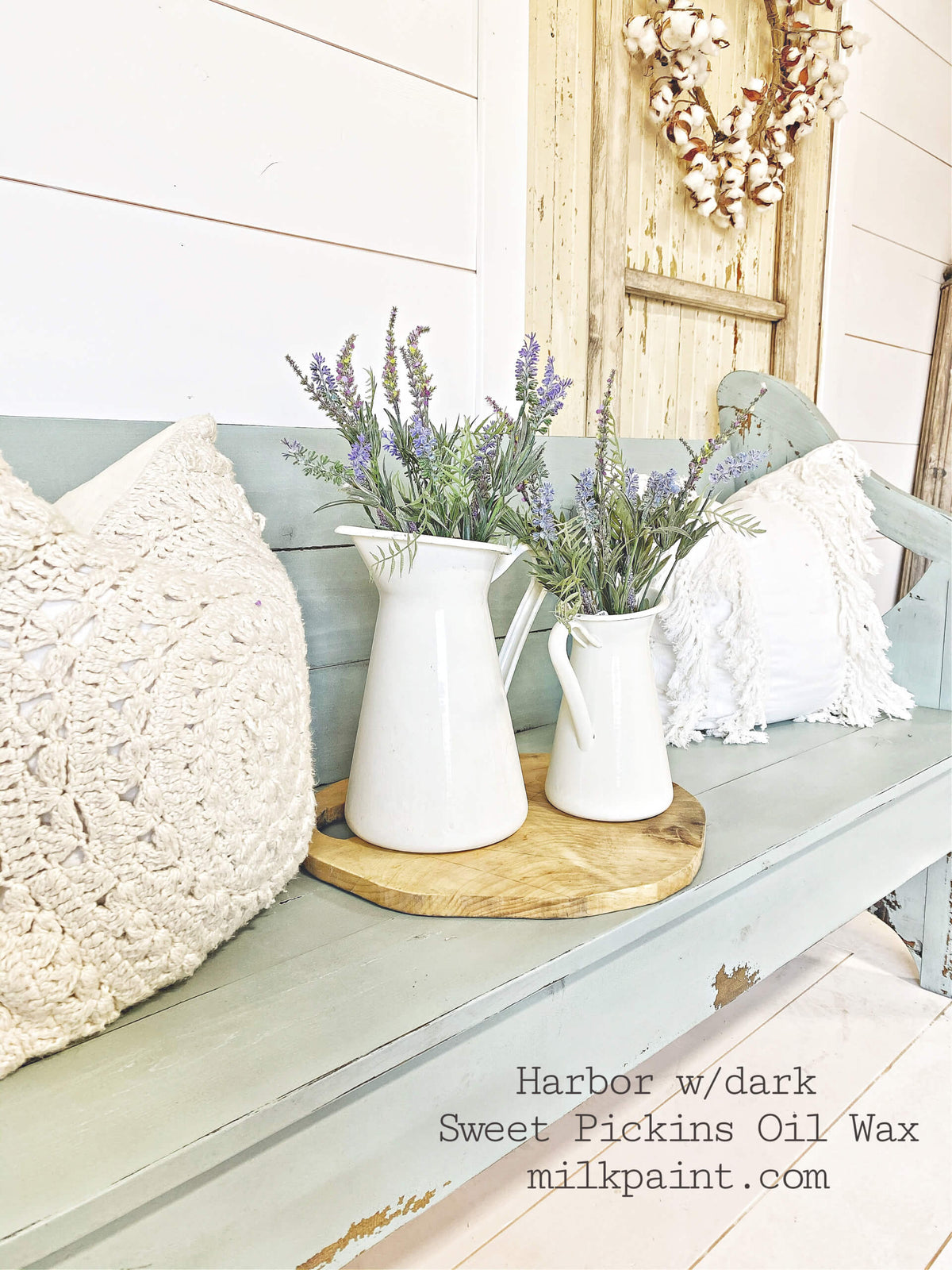 A rustic painted bench in a soft sage green color, styled with two white ceramic pitchers filled with lavender on a wooden tray, textured cream pillows, and a white shawl on a porch.