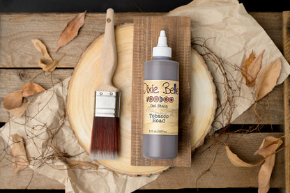 Bottle of Dixie Belle Gel Stain Tobacco Road with a brush on a wooden surface with leaves.