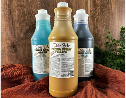 Three bottles of Dixie Belle Patina Spray in different colors on a wooden surface with a red fabric and greenery.