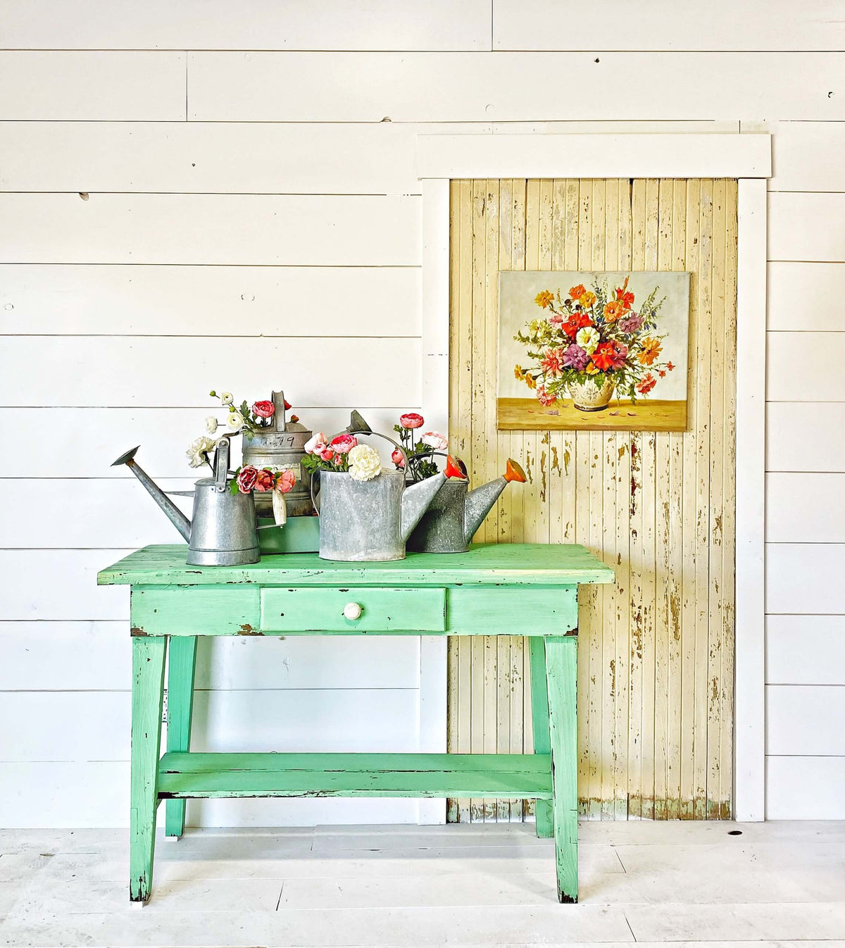 A distressed mint green wooden table styled with vintage galvanized metal watering cans and small flower arrangements, set against a white shiplap wall and a rustic weathered wood door with a floral painting hanging above.