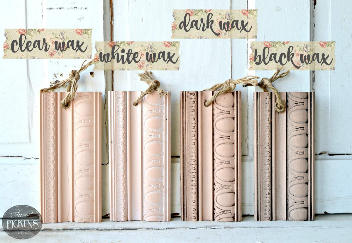 Five decorative book spine ornaments in blush pink, peach, and beige tones are arranged against a white distressed wood background. Above them are three paper labels reading 'clear wax,' 'white wax,' and 'black wax,' each tied to one ornament with twine.