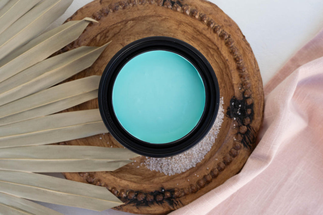 Lani's Lagoon Terra Clay Paint, placed on a rustic wooden slice with dried palm leaves and pale pink fabric around it.