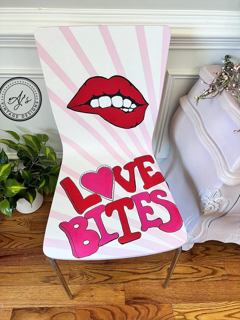 A white accent chair with pink sunburst stripes, featuring a graphic of red lips and the text 'LOVE BITES' in bold pink and red letters, styled in a home setting on a wooden floor.