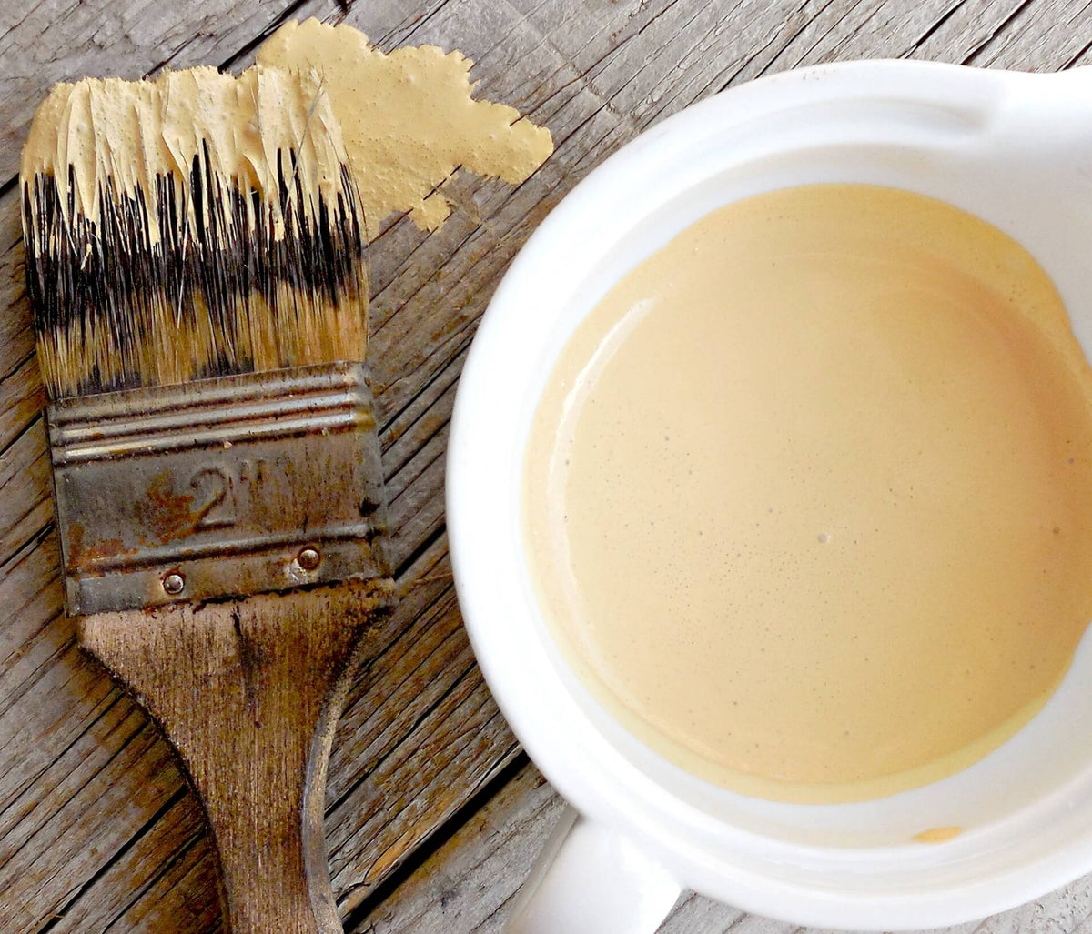 Wooden brush with paint next to a container of white paint on a wooden surface
