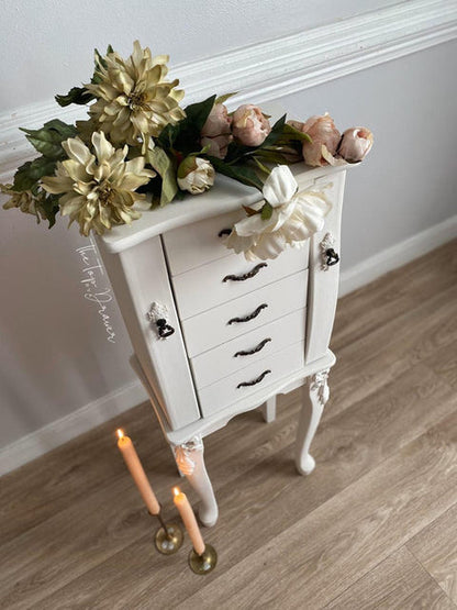 A white vintage-style jewelry chest with curved legs and black drawer pulls, styled with faux flowers on top and candles on the floor in a home setting.
