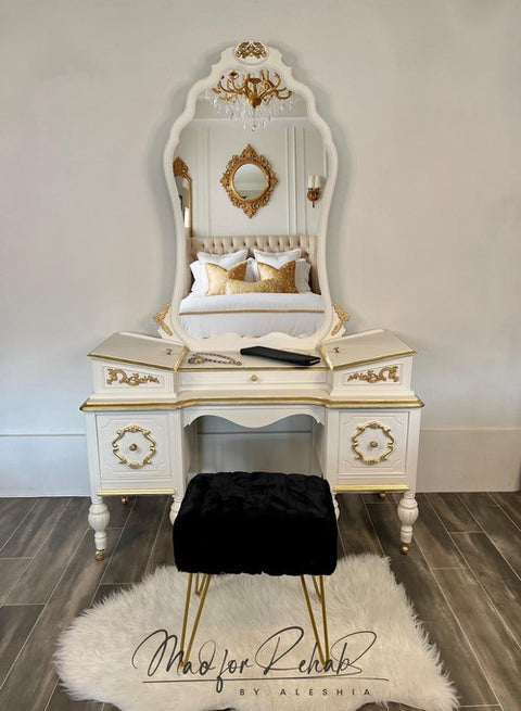 An ornate white and gold vanity table with a large arched mirror, styled with white and gold pillows, a black velvet stool, and a white faux fur rug on a wood floor.